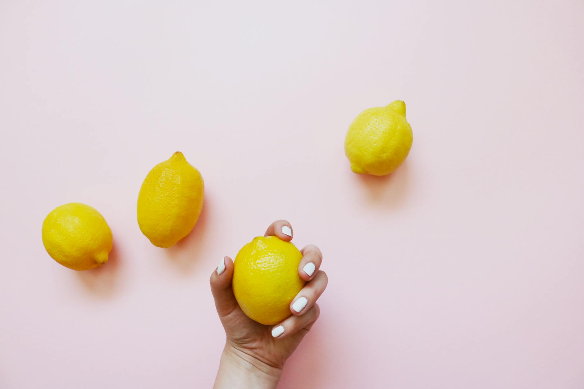Una mano con unghie bianche che tiene un limone su uno sfondo rosa tenue, circondata da tre limoni aggiuntivi.