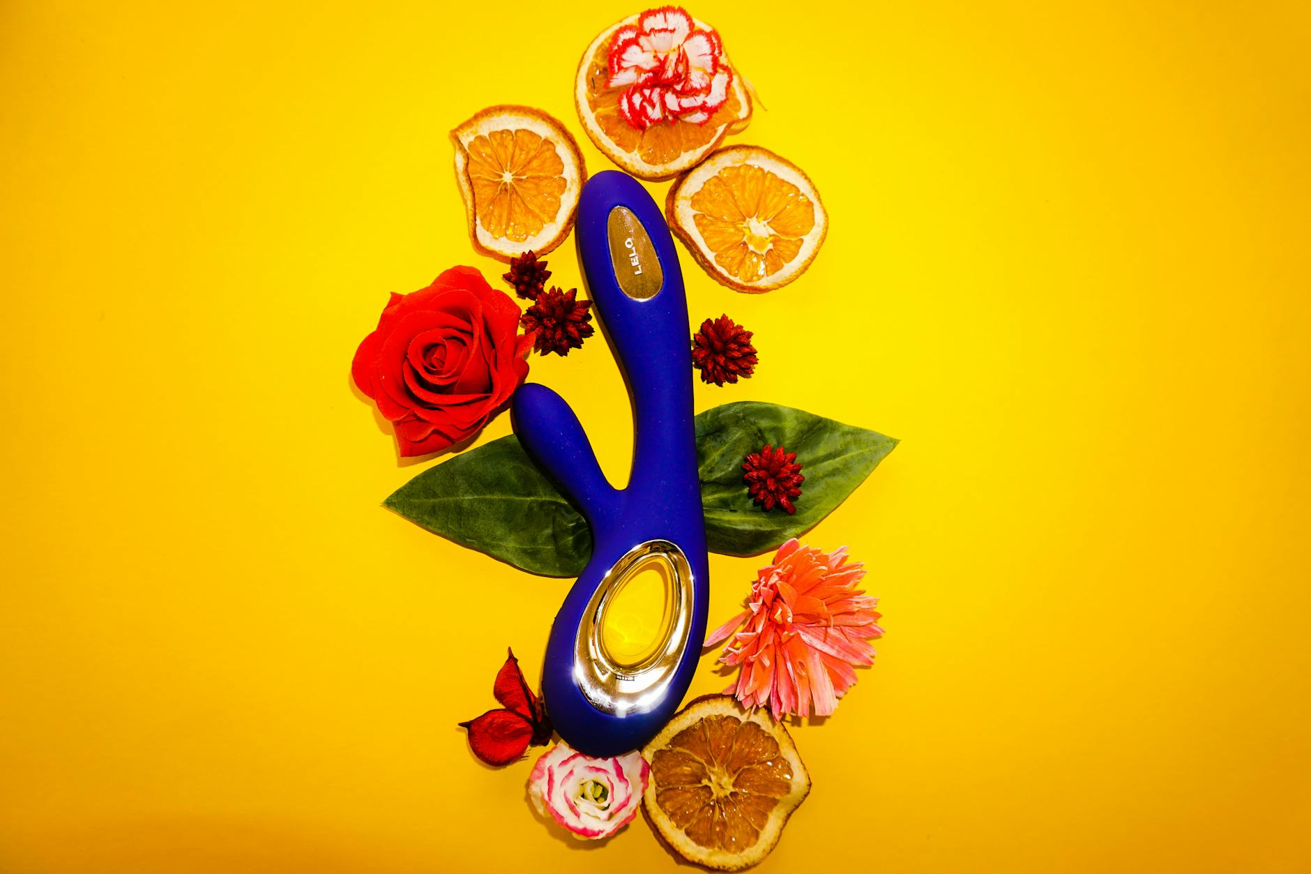 Un vibratore blu circondato da fiori e fettine d'arancia su sfondo giallo